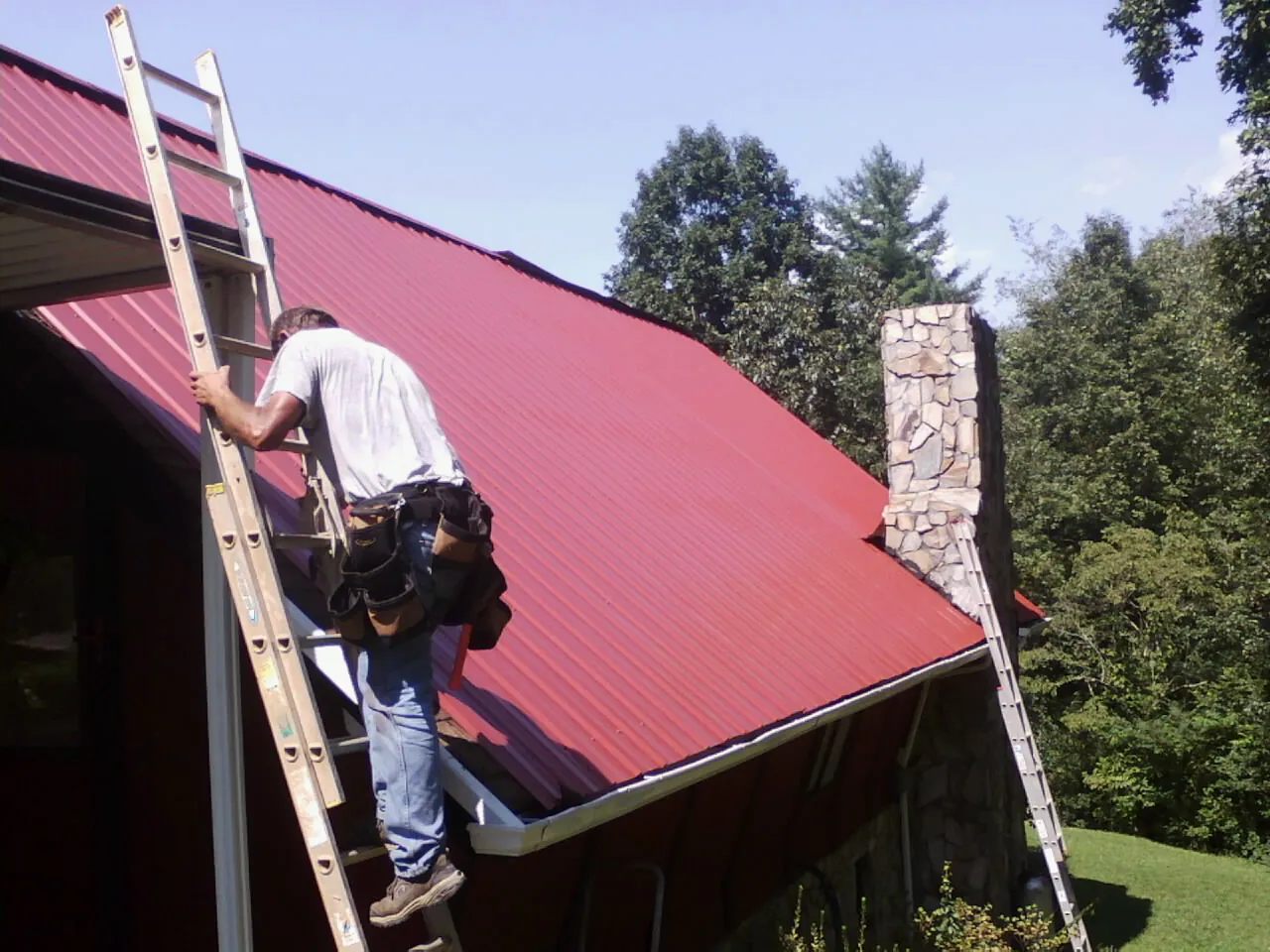Roofer on a ladder beside a red metal roof and stone chimney — Soffit & Fascia Repair by Roofing Brothers in Scotts Mills